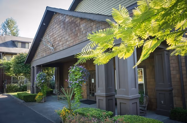 Exterior view of Weatherly Inn Lake Meridian showing the entrance with large brown pillars, hanging flower baskets, and surrounding greenery including bushes and trees.