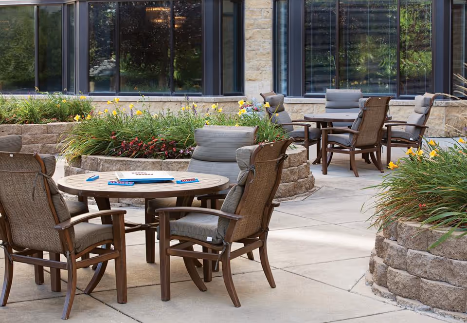 Outdoor patio area with round tables and cushioned chairs arranged around them. Raised stone flower beds with green plants and yellow flowers surround the seating area. Large windows of a building are visible in the background.
