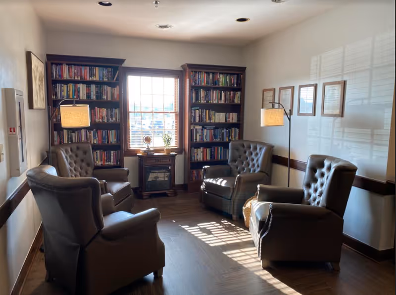 Sunlit reading room with four leather armchairs arranged around a window flanked by bookshelves and floor lamps.