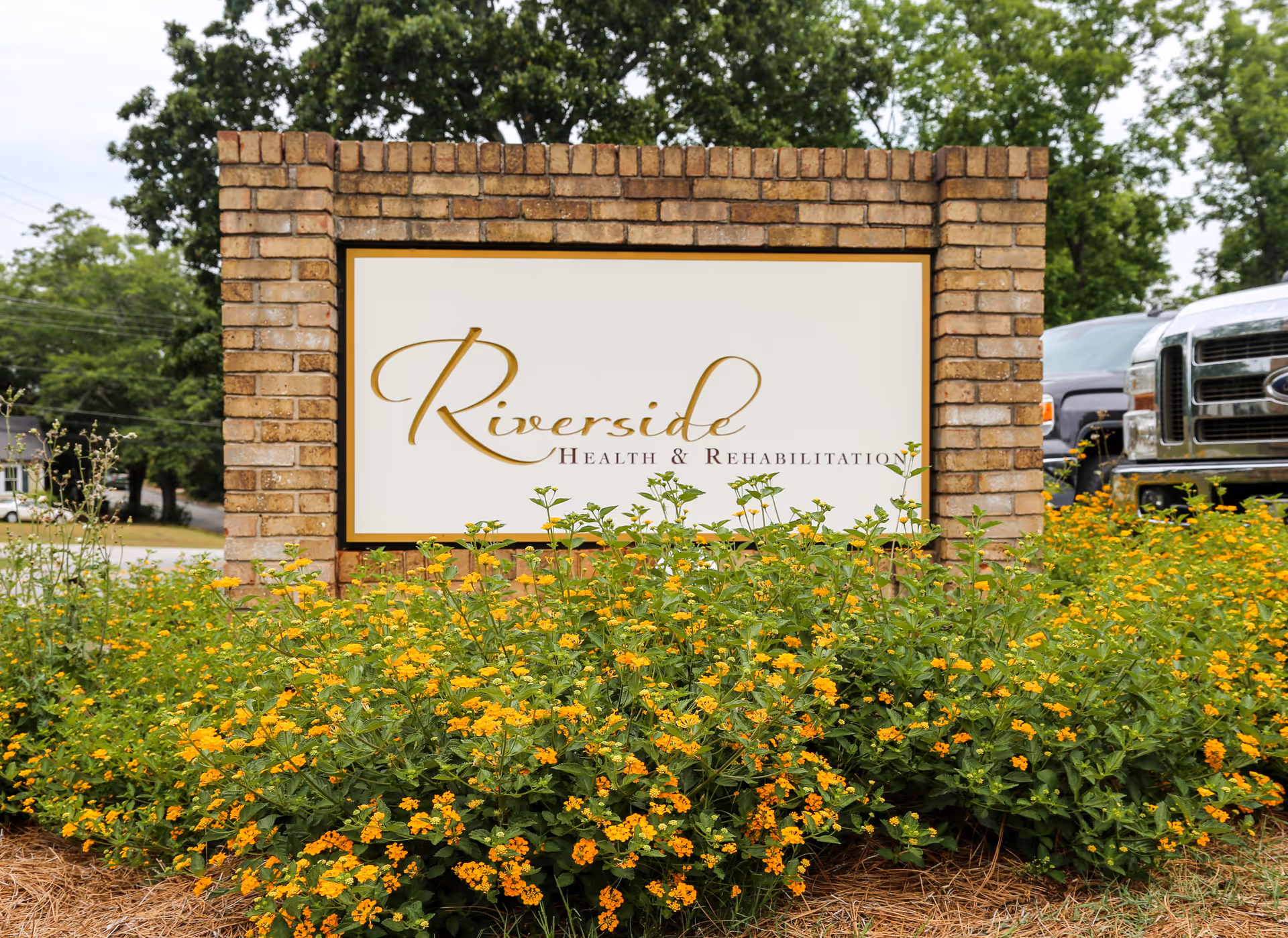 A brick sign for Riverside Health & Rehabilitation surrounded by yellow flowering plants with trees and parked vehicles in the background.