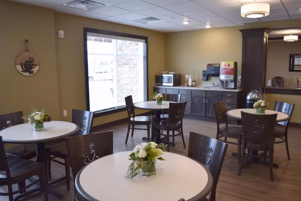 A small dining area with round tables and wooden chairs, each table decorated with a small flower arrangement. The room has a large window with blinds, a counter with a microwave, a beverage dispenser, and a sink. The walls are painted in a warm beige color and the floor has wood-style flooring.