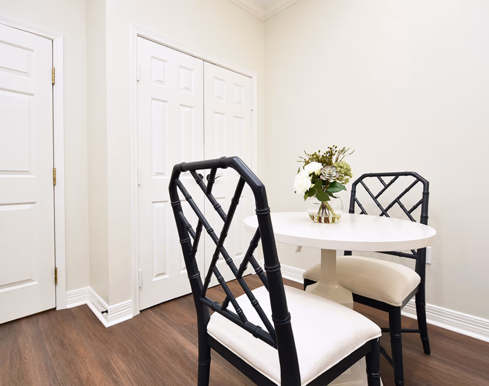 Small dining area with a round white table, two black chairs with white cushions, and a vase of flowers against light walls and wood flooring.