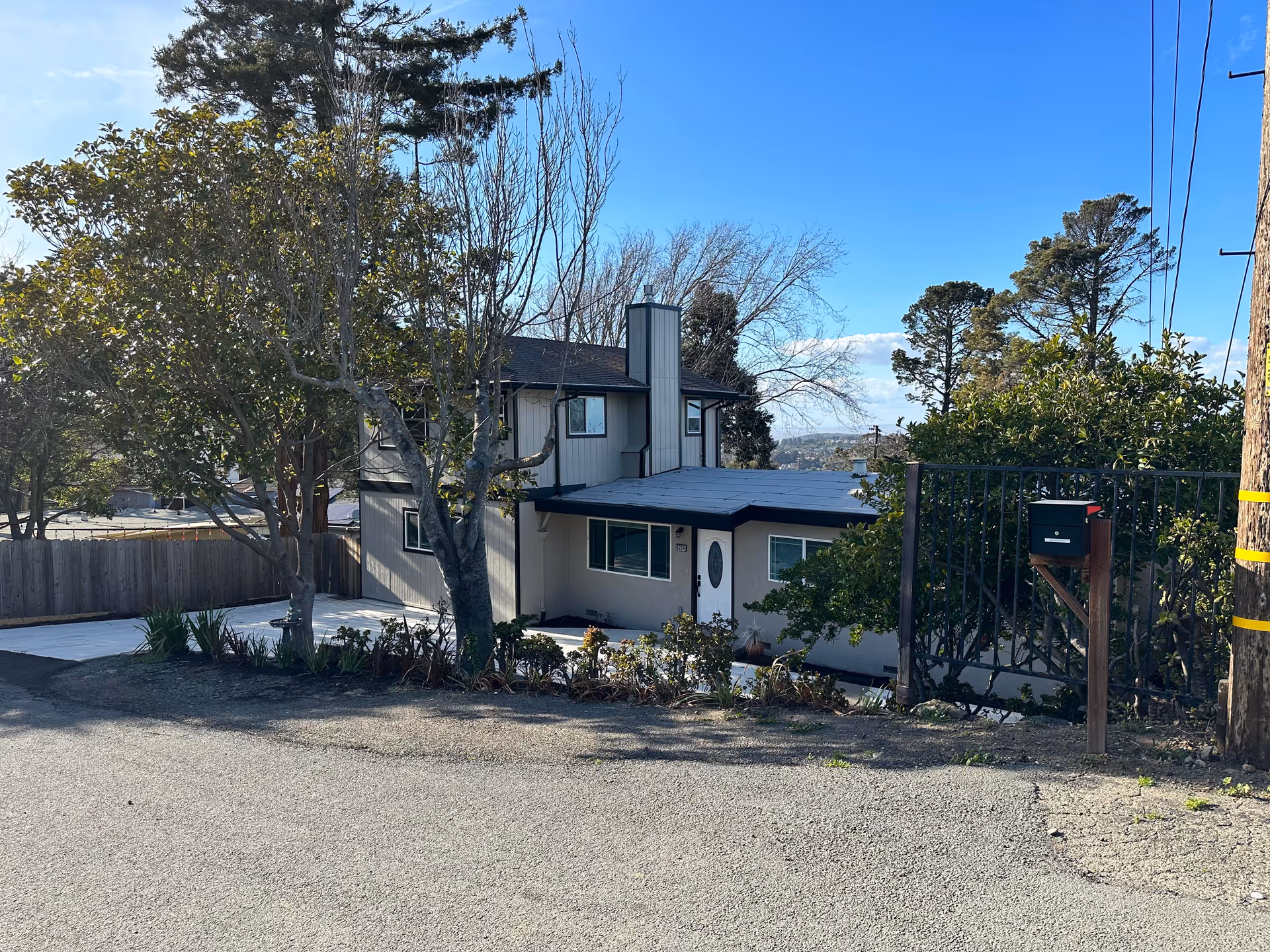 Exterior view of a two-story residential building with light-colored walls and a dark roof, surrounded by trees and bushes. A black metal gate and a mailbox are visible on the right side, with a clear blue sky in the background.