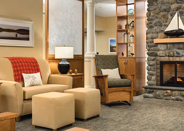 Cozy living room area with a beige sofa draped with a red plaid throw, a patterned armchair with a white pillow, two beige ottomans, a black table lamp on a wooden side table, a stone fireplace with a small sailboat model on the mantel, and wooden shelving with decorative items.