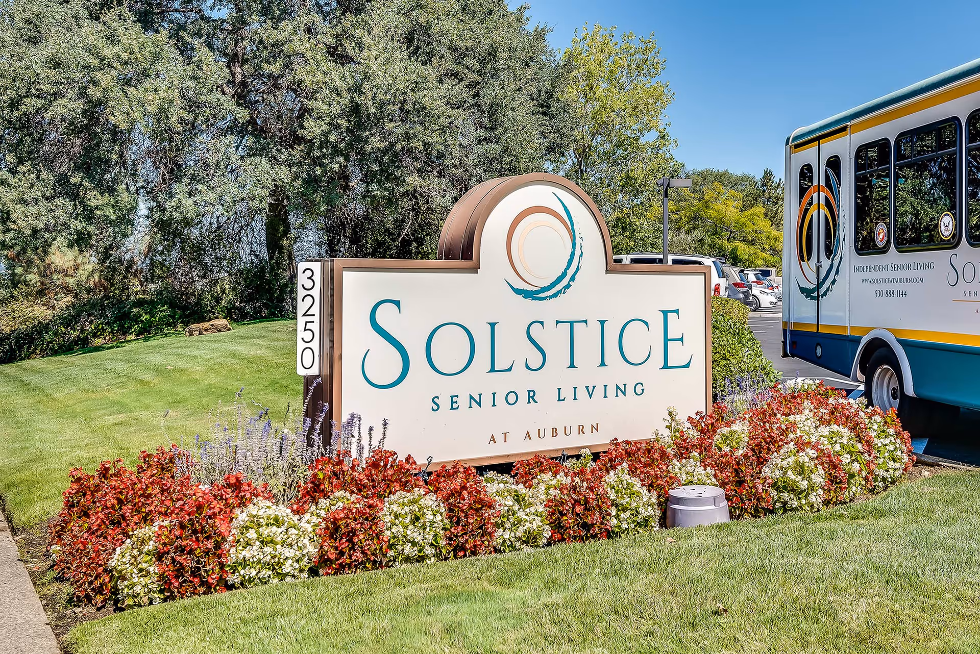 Outdoor view of the entrance sign for Solstice Senior Living at Auburn, surrounded by colorful flowers and green grass, with trees and a shuttle bus in the background.