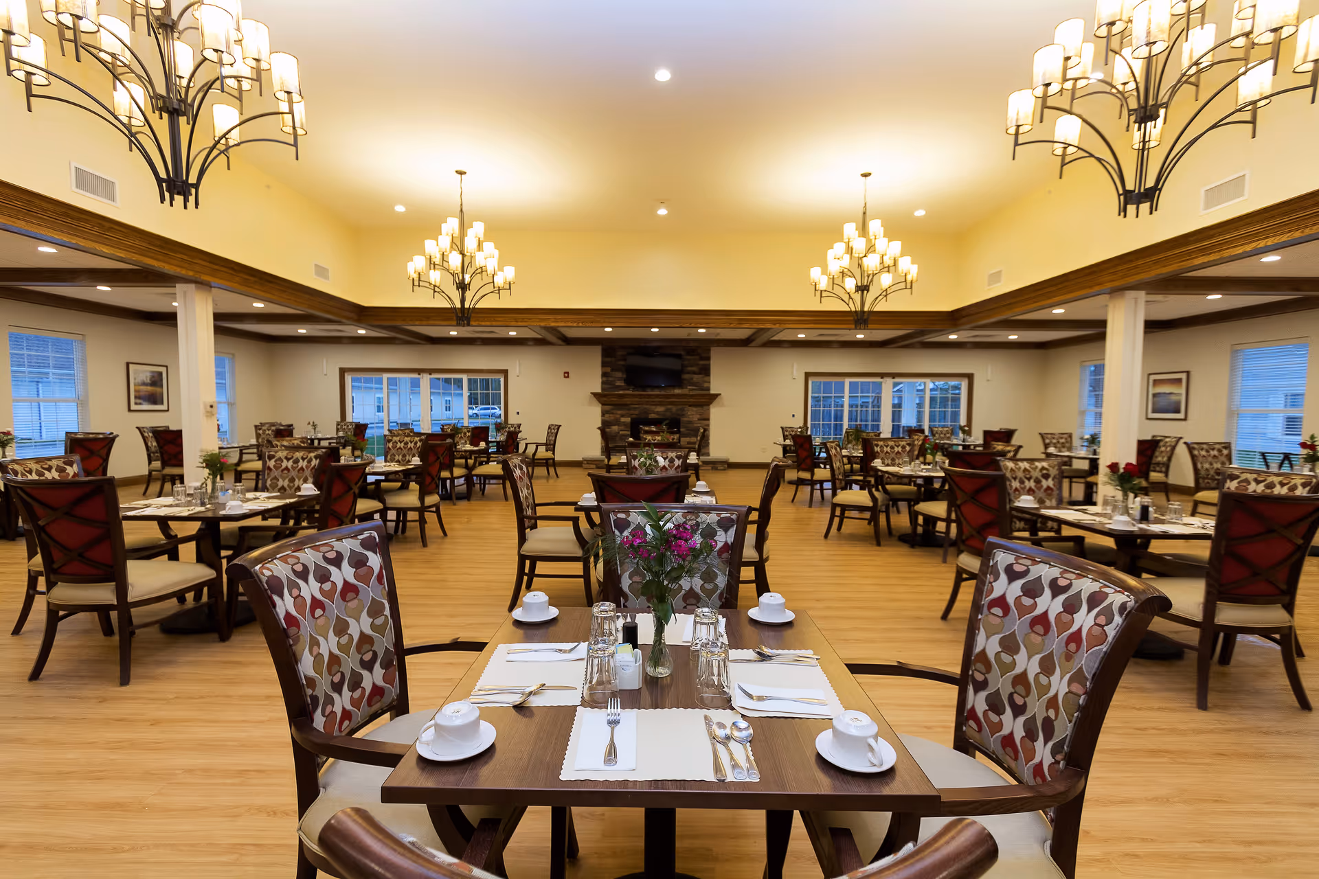 A spacious, well-lit dining room with multiple set tables, patterned chairs, chandeliers, and a fireplace at the far wall.