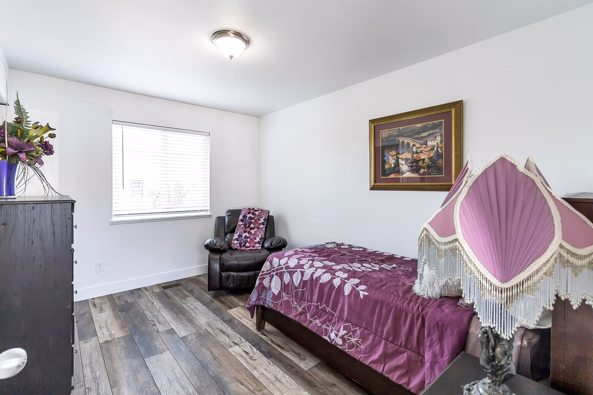 Small bedroom with purple bedding, dresser, chair, and framed wall art.