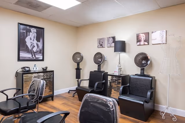 Interior of a salon area with three black salon chairs, two hair drying stations, a small mirrored cabinet with a lamp and tissue box on top, a vintage black rotary phone on another mirrored cabinet, and black and white photos of Marilyn Monroe on the walls. The floor is wooden and the walls are beige.