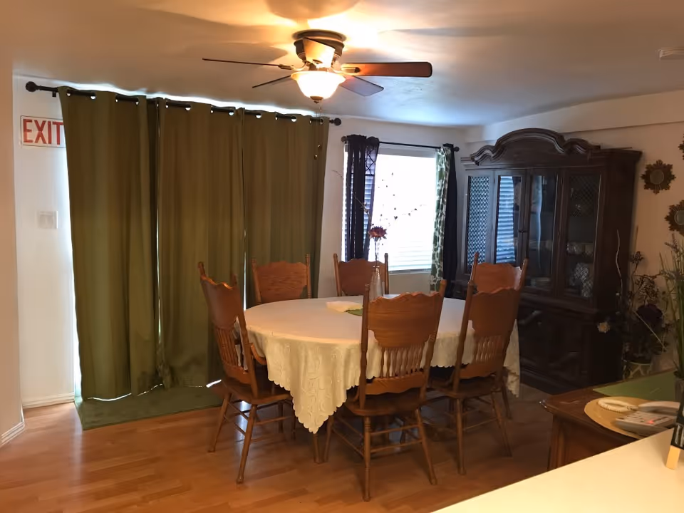 A dining room with a round table covered by a tablecloth surrounded by six wooden chairs, a china cabinet, green curtains, and a ceiling fan.