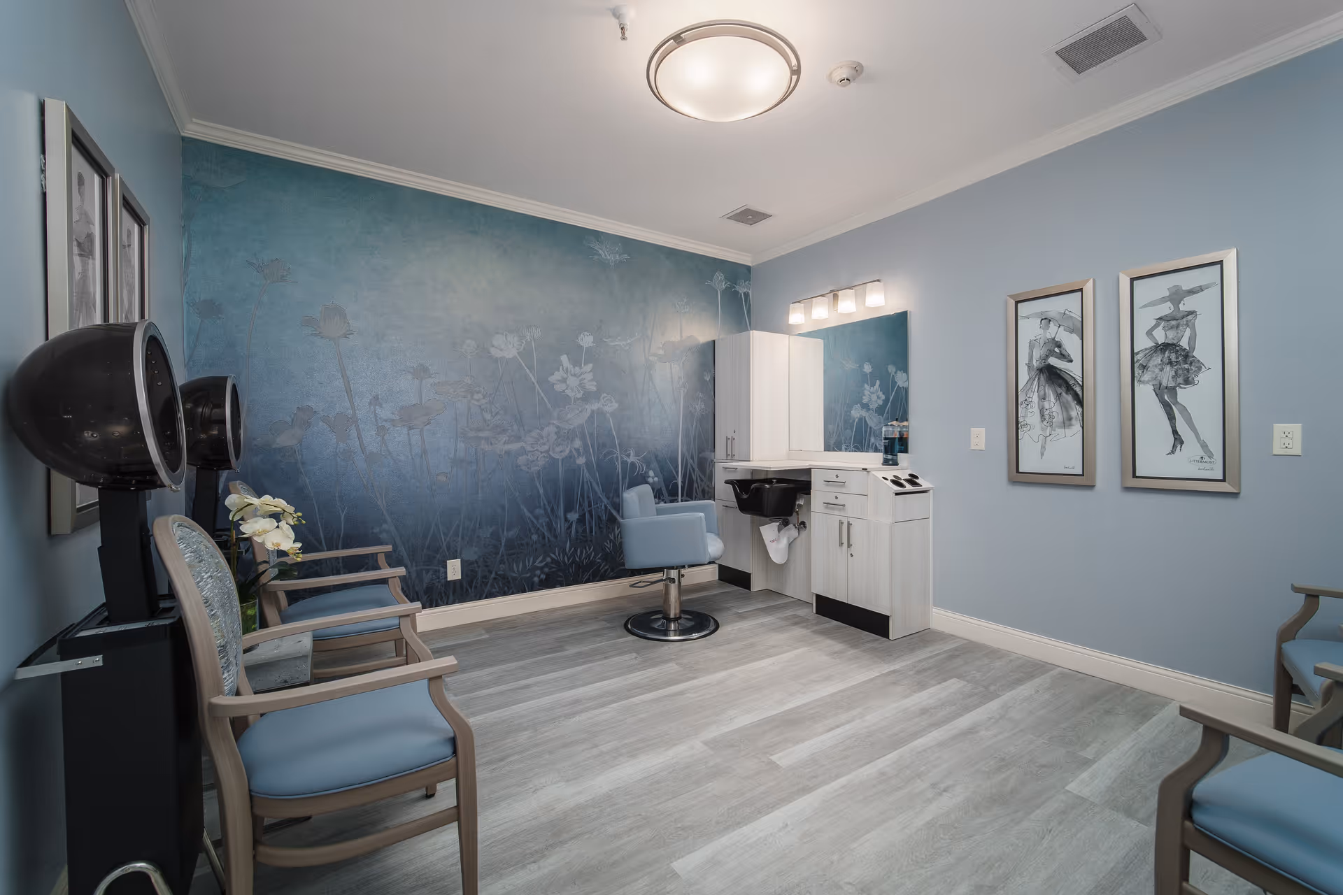 A small salon room with light blue walls and a floral mural on one wall. The room features a salon chair in front of a white cabinet with a mirror and a wash basin. There are two black hair dryers on stands and several blue cushioned chairs along the walls. The floor is light wood, and the room is softly lit by a ceiling light and a wall-mounted light fixture above the mirror. Two framed fashion sketches hang on one wall.