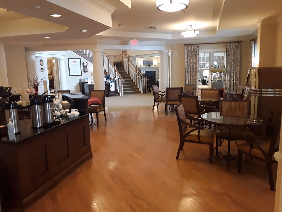 Interior view of a senior living facility common area with wooden floors, round tables with chairs, a coffee station with multiple coffee dispensers, and a staircase leading to an upper floor. The space is warmly lit with ceiling lights and decorated with curtains and framed pictures.