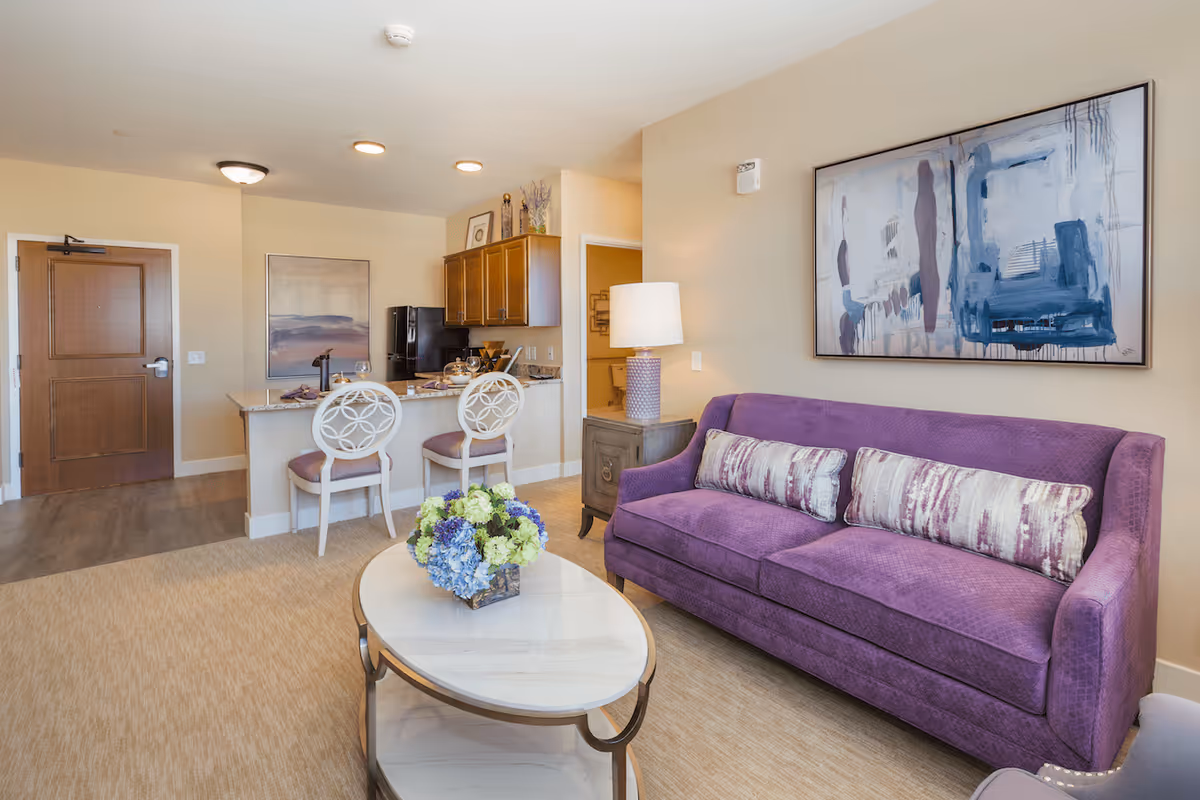 A cozy living room area with a purple sofa adorned with two patterned cushions, a marble-top coffee table with a floral arrangement, and a side table with a lamp. In the background, there is a kitchen counter with two white chairs, wooden cabinets, and a refrigerator. The walls are decorated with abstract paintings, and the entrance door is visible on the left.