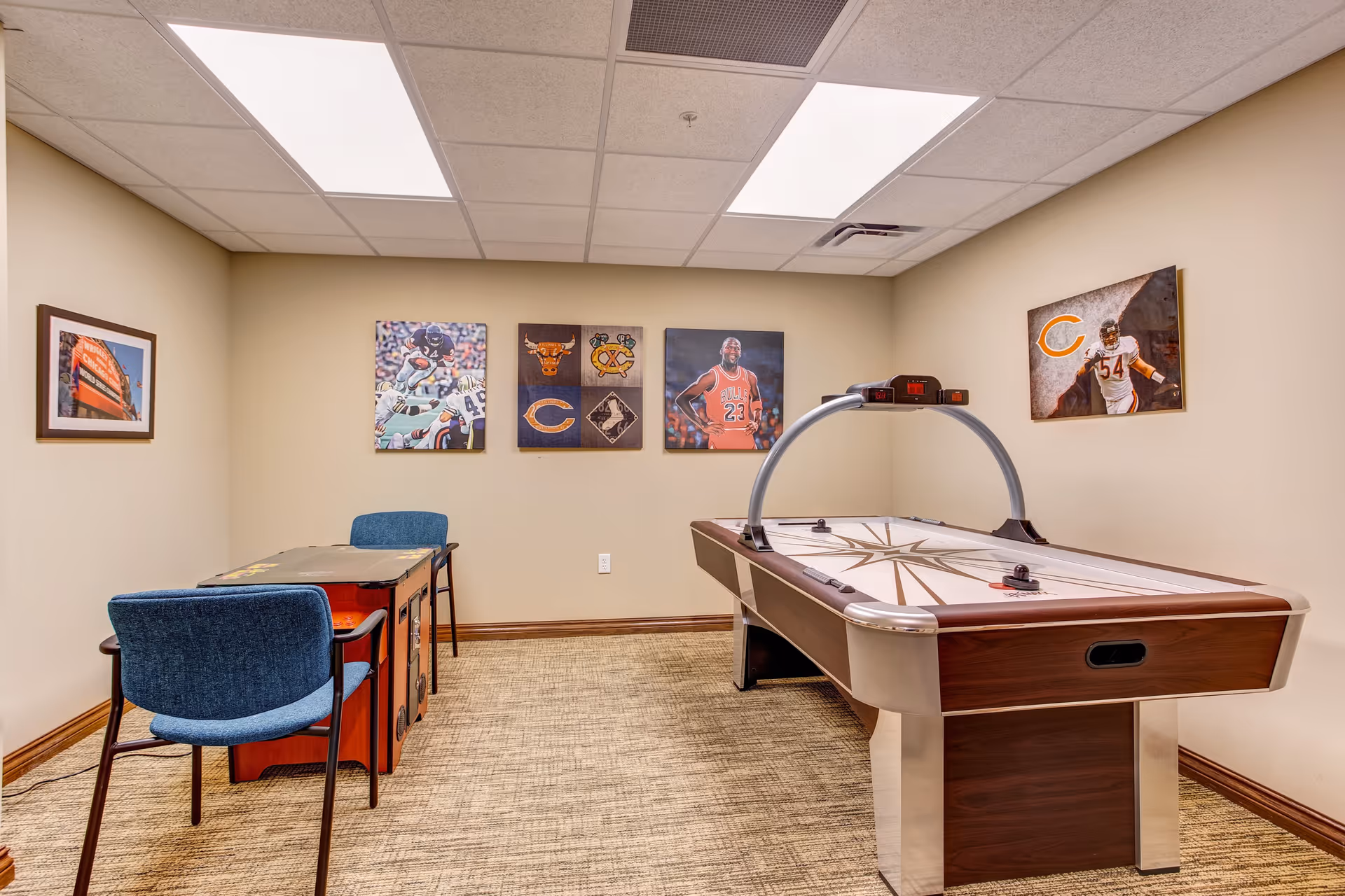 A recreation room with an air hockey table and a small table with two blue chairs. The walls are decorated with framed sports-themed pictures, including images of football players and basketball player Michael Jordan. The room has beige walls, a carpeted floor, and a drop ceiling with fluorescent lights.