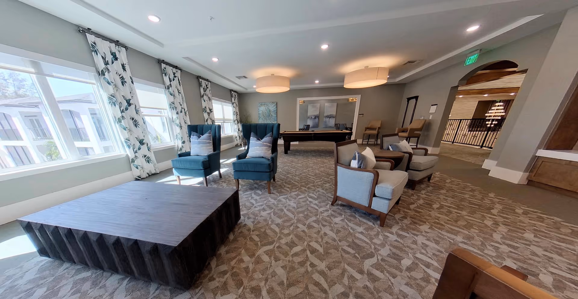 A spacious and well-lit common area with large windows covered by floral curtains. The room features comfortable seating including two blue armchairs with pillows, two beige armchairs with pillows, and a large dark wooden coffee table. In the background, there is a pool table and additional chairs along the wall. The carpet has a leaf pattern, and the ceiling has modern round light fixtures. An open doorway leads to another area with a fireplace and more seating.