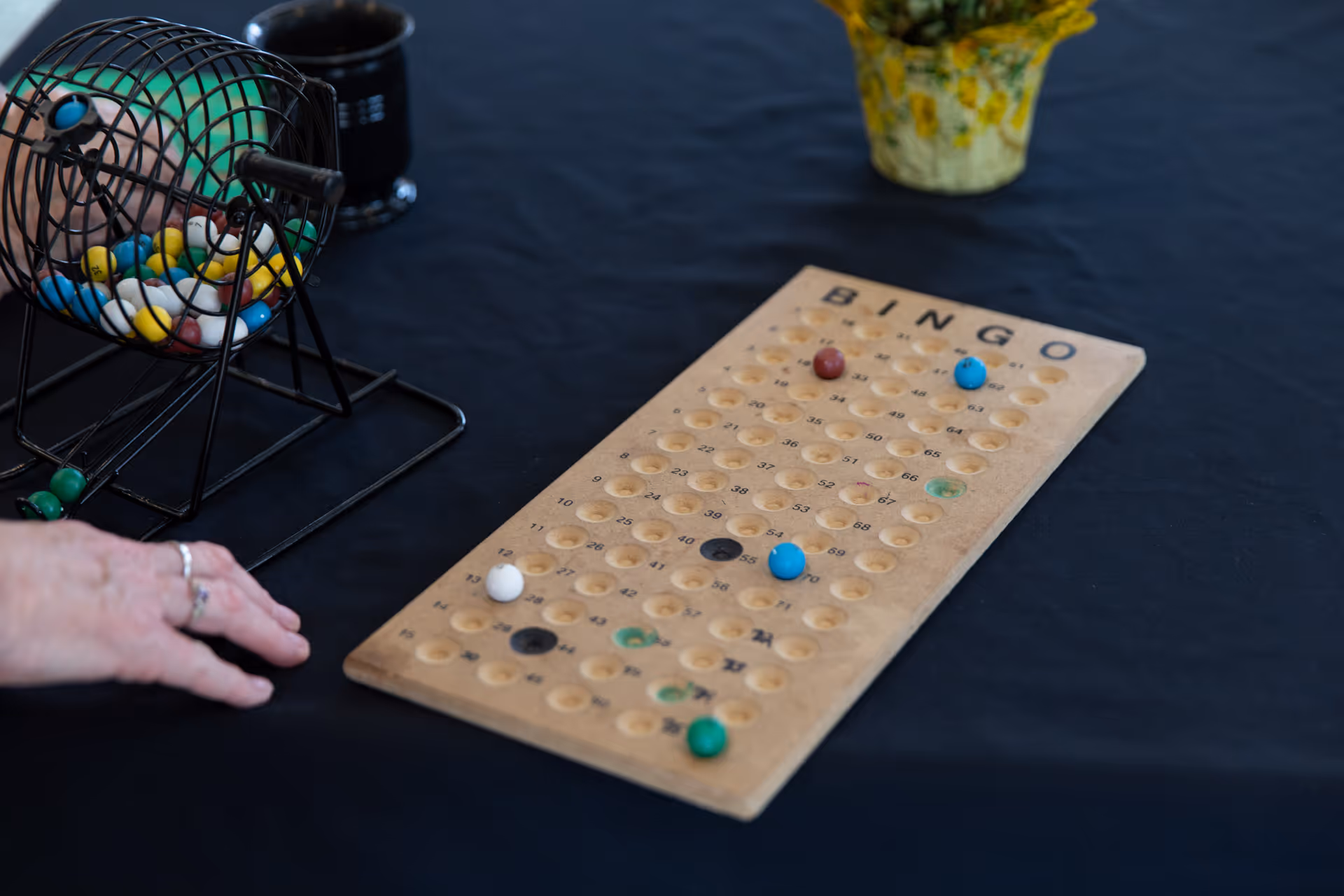 A close-up view of a bingo game setup on a black tablecloth, featuring a wooden bingo board with colored markers placed on it, a metal cage with bingo balls inside, and a person's hand resting on the table near the cage.