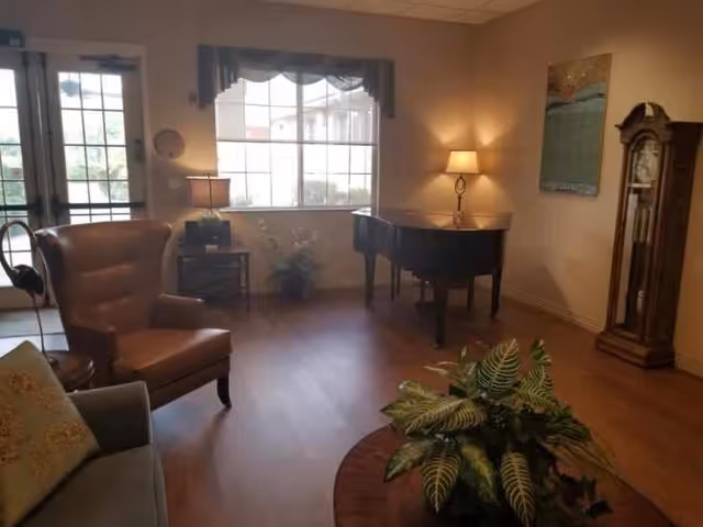 A cozy living room with wooden flooring featuring a brown leather armchair, a gray sofa with a decorative pillow, a round wooden coffee table with a green leafy plant, a grand piano with a table lamp on it, a tall grandfather clock, and a large window with a valance letting in natural light.