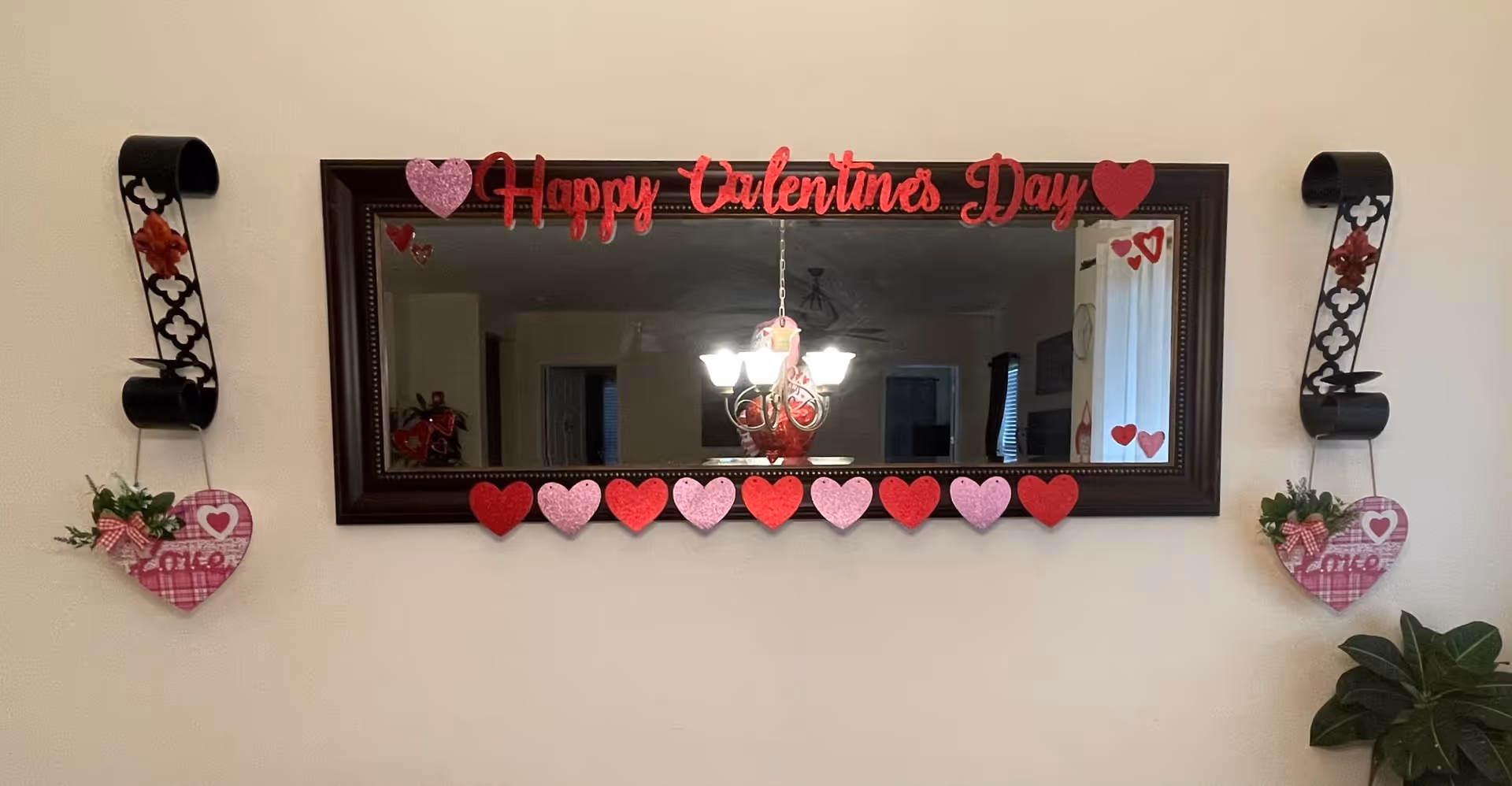 A rectangular wall mirror with a dark wooden frame decorated with red and pink hearts and the words 'Happy Valentine's Day' in red glittery letters. On either side of the mirror are black metal wall sconces holding heart-shaped decorations with the word 'Love' and small plants. A chandelier with multiple lights is reflected in the mirror.
