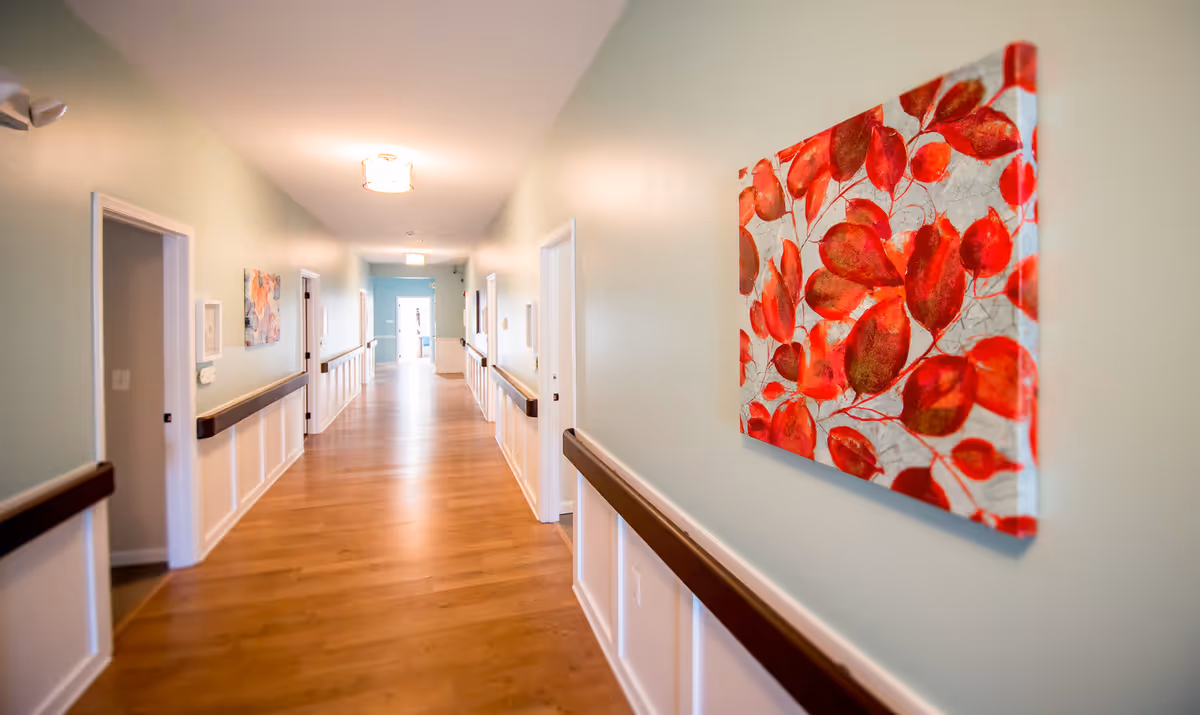 A well-lit hallway in a senior living facility with light green walls, wooden flooring, and handrails on both sides. Several doorways line the corridor, and colorful artwork featuring red leaves is displayed on the right wall.