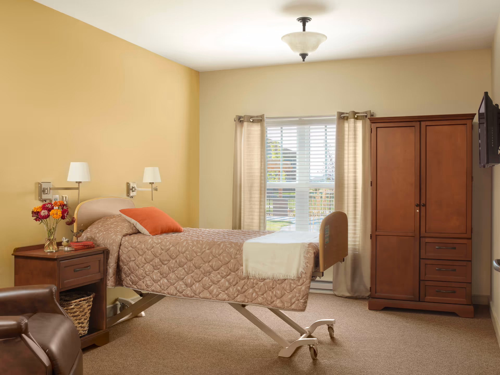 A neatly arranged bedroom in a senior living facility with a single adjustable bed covered in a beige quilt and an orange pillow. There is a wooden nightstand with a vase of flowers and books, a brown leather chair, a wooden wardrobe, and a wall-mounted TV. A window with beige curtains allows natural light into the room.