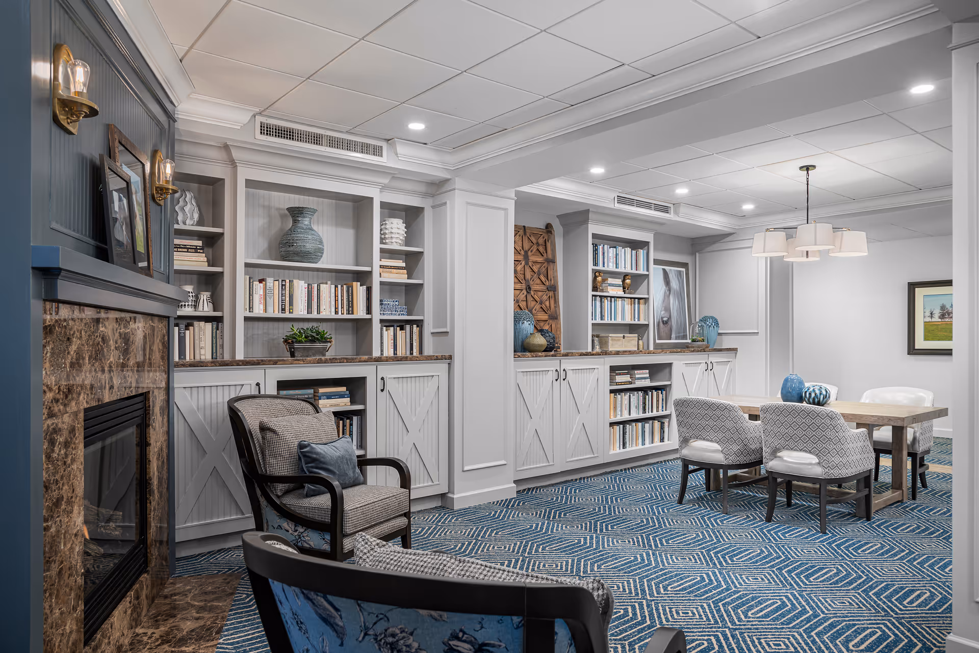 A cozy and well-lit common area featuring built-in bookshelves filled with books and decorative vases, a marble fireplace with framed pictures and wall sconces above it, a wooden table with four upholstered chairs, and a blue patterned carpet covering the floor.