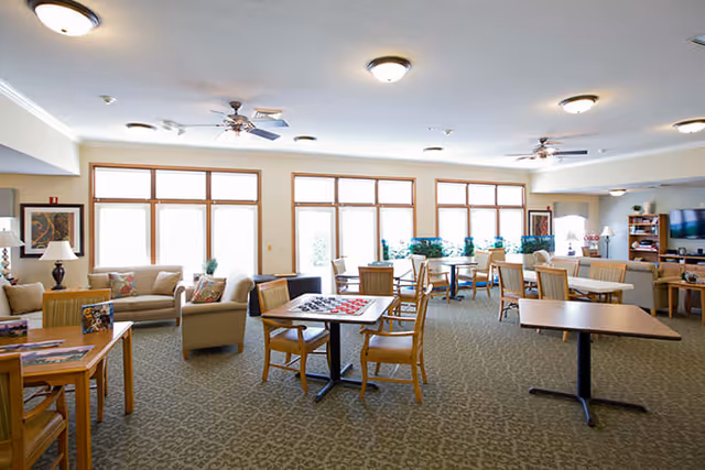 A spacious, well-lit common area with multiple tables and chairs arranged for socializing or activities. There are large windows letting in natural light, ceiling fans, and comfortable seating including sofas and armchairs. The room has a carpeted floor and neutral-colored walls with some framed artwork and plants near the windows.