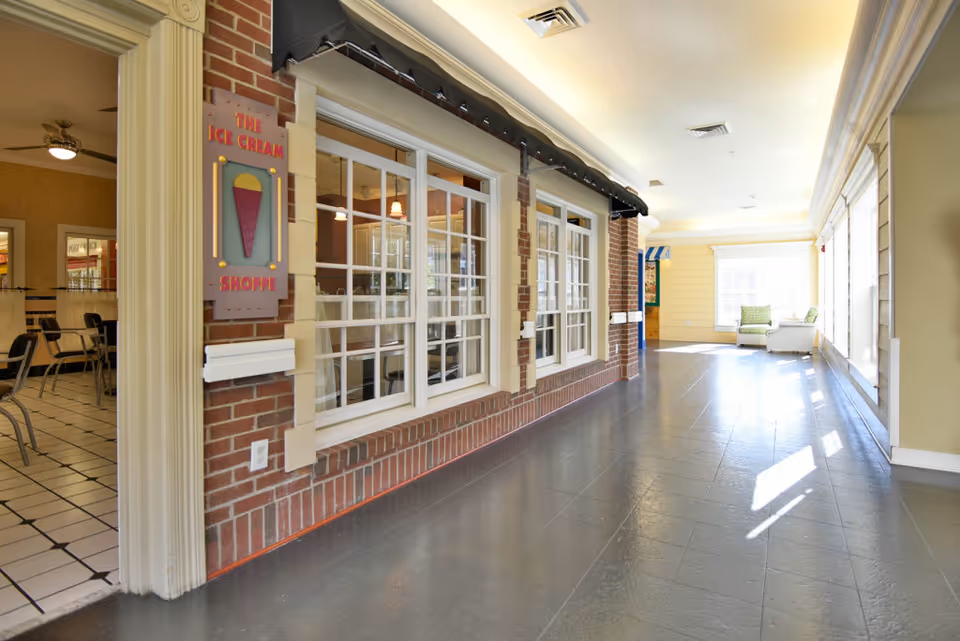 Interior hallway of a senior living facility with a brick wall featuring large windows and a sign that reads 'The Ice Cream Shoppe'. To the left, there is a dining area with tables and chairs visible through an open doorway. The hallway is well-lit with natural light coming from large windows on the right side, and there are two chairs and a small table near the far end.