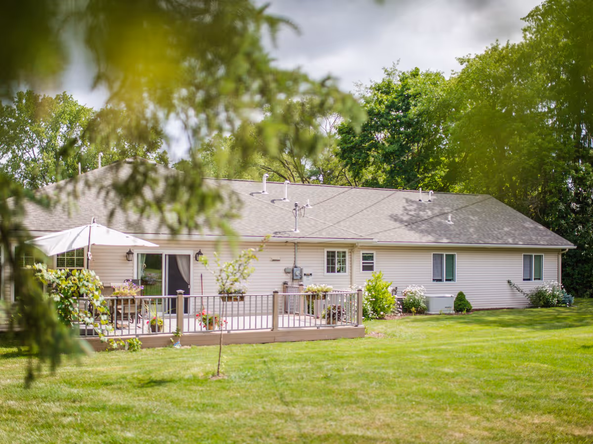 View of a single-story beige building with a gray roof, surrounded by green grass and trees. The building has a wooden deck with potted plants and a white patio umbrella. The scene is framed by blurred green leaves in the foreground.