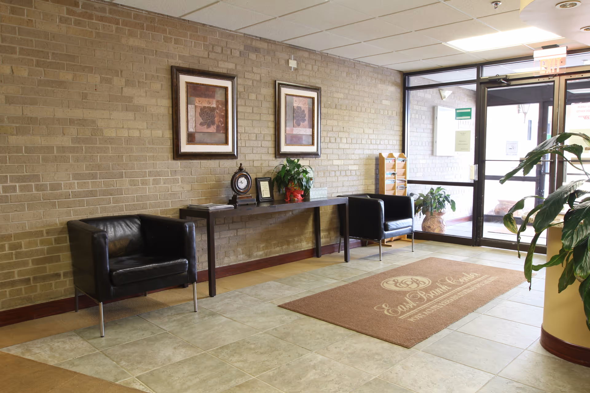 A lobby area with two black leather chairs on either side of a long wooden table against a brick wall. The table holds a clock, a framed certificate, and a plant. Two framed artworks hang above the table. There is a glass door entrance with plants near it and a brown floor mat with the text 'East Bank Center' and a logo.