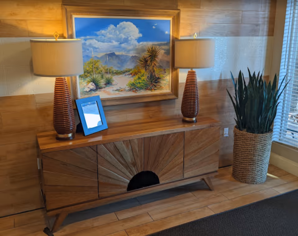 Wooden console table topped with two lamps, a small digital frame and a framed desert painting on a wood-paneled wall, with a tall potted plant to the right.