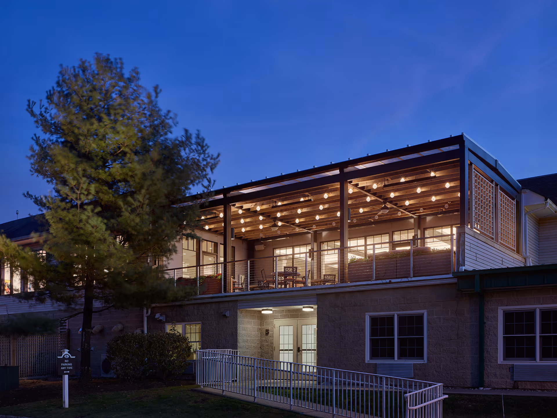 Exterior view of a senior living facility at dusk featuring a well-lit covered balcony with string lights and outdoor seating. The building has large windows and a ramp leading to a door on the ground level. A tree and some shrubs are visible in the foreground.