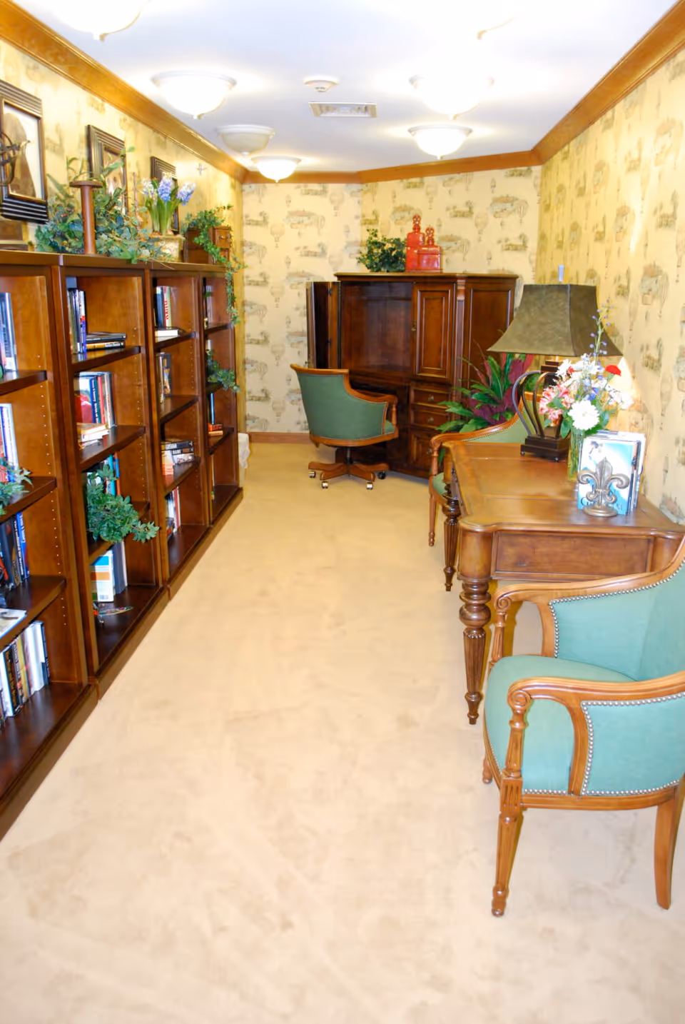 A cozy interior room with beige carpet and wallpaper featuring a vintage hot air balloon pattern. On the left side, there are wooden bookshelves filled with books and decorated with green plants. On the right side, there is a wooden desk with a lamp and a flower arrangement, accompanied by green upholstered chairs with wooden frames. At the far end of the room, a wooden cabinet and a green swivel chair are visible.