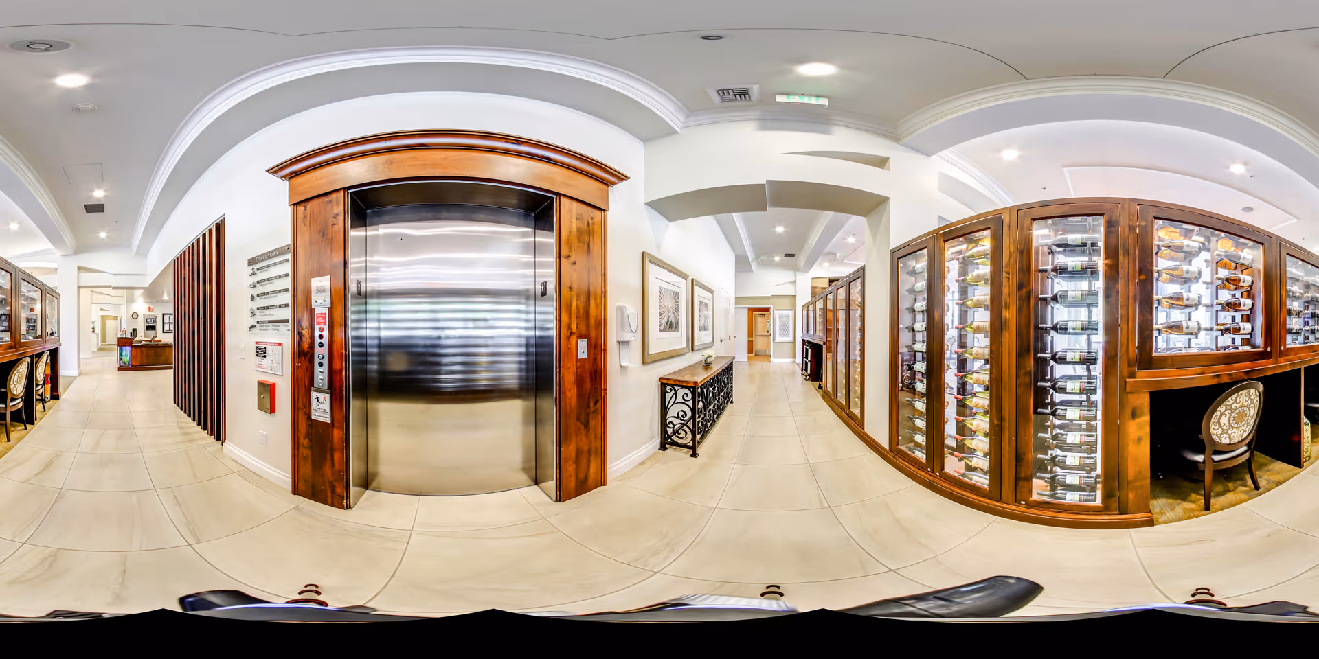 Interior hallway of a senior living facility featuring a stainless steel elevator with a wooden frame, a decorative console table with framed artwork on the wall, and a large glass-enclosed wine rack with numerous bottles. The space has tiled floors, recessed lighting, and a clean, modern design.