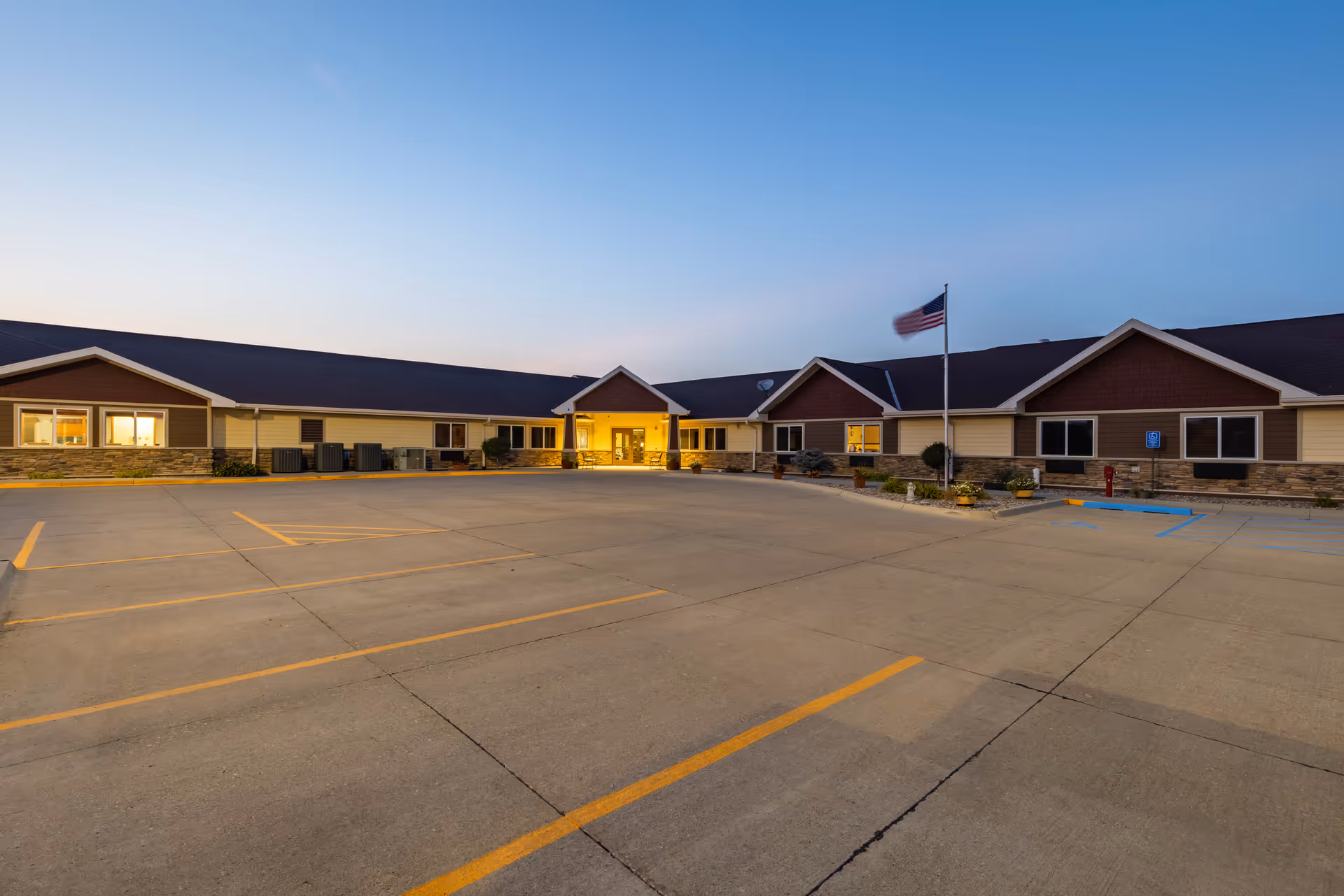 Exterior view of a single-story senior living facility building at dusk with a large empty parking lot in front, an American flag on a flagpole, and warm lights glowing from the entrance and windows.