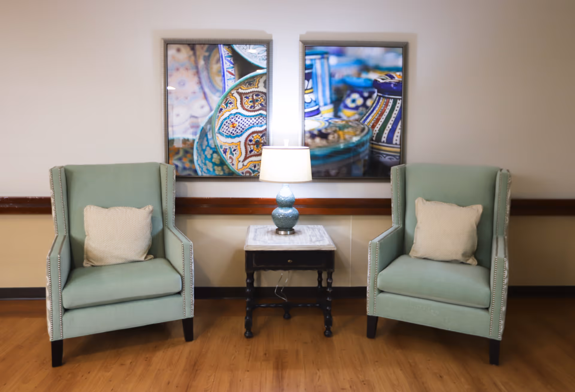 Two light green upholstered armchairs with beige cushions are positioned on either side of a small wooden side table with a marble top. A blue ceramic table lamp with a white shade sits on the table. Behind the chairs are two framed colorful abstract art prints mounted on a beige wall. The floor is wooden.