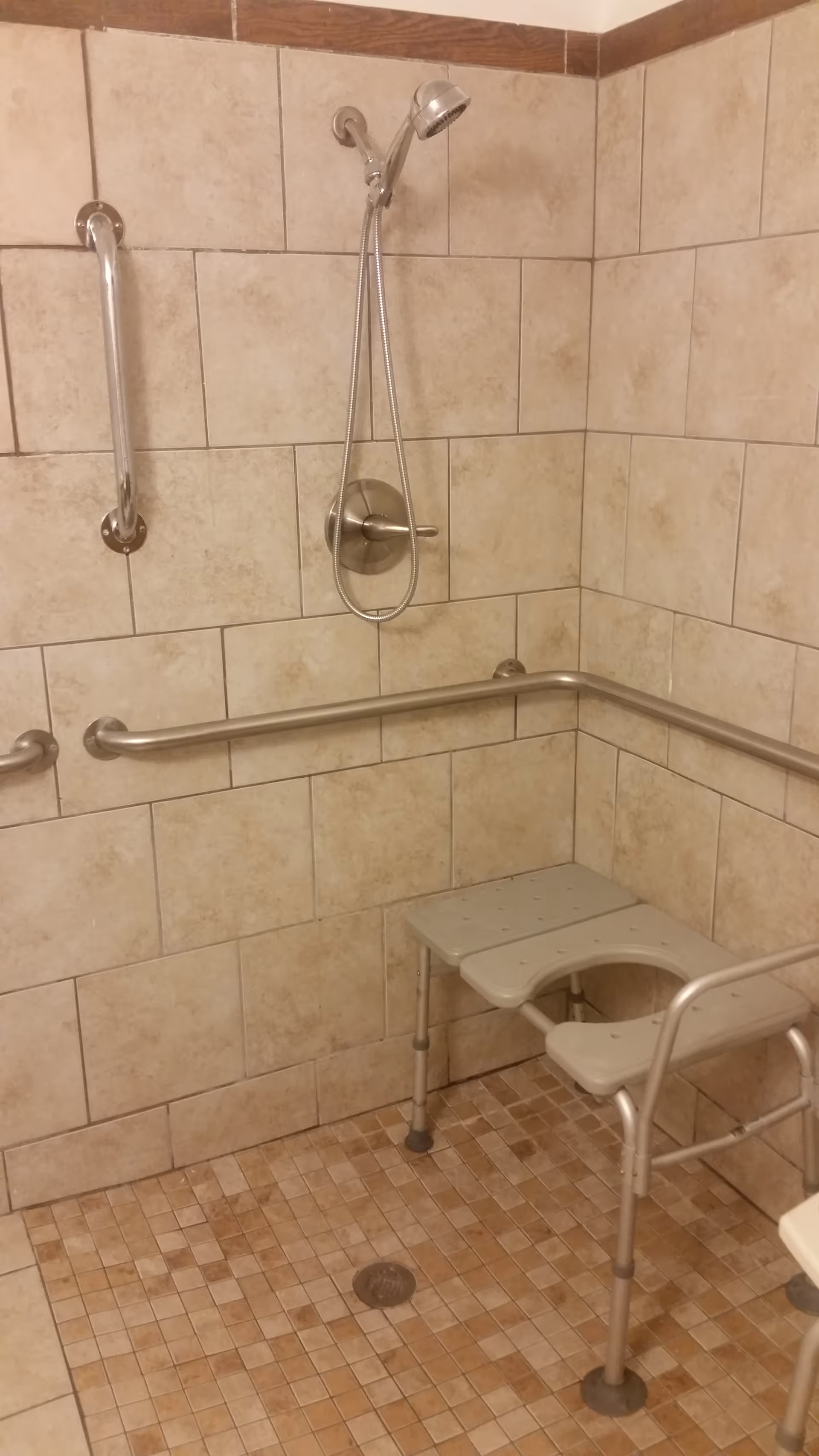 Accessible shower area with beige tiled walls and floor, equipped with a handheld showerhead, multiple stainless steel grab bars, and a gray shower chair with a cut-out seat.