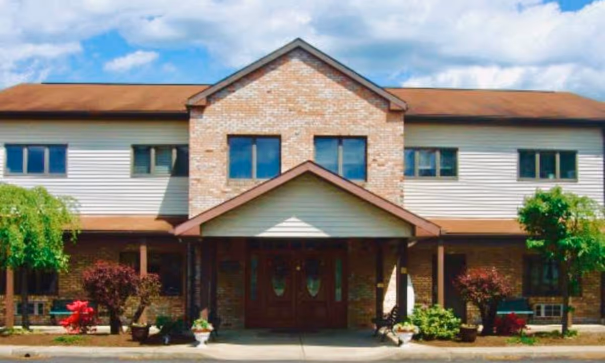 Front exterior view of a two-story building with a brick and siding facade, a peaked roof, and a covered entrance. There are small trees and shrubs planted in front of the building under a partly cloudy sky.