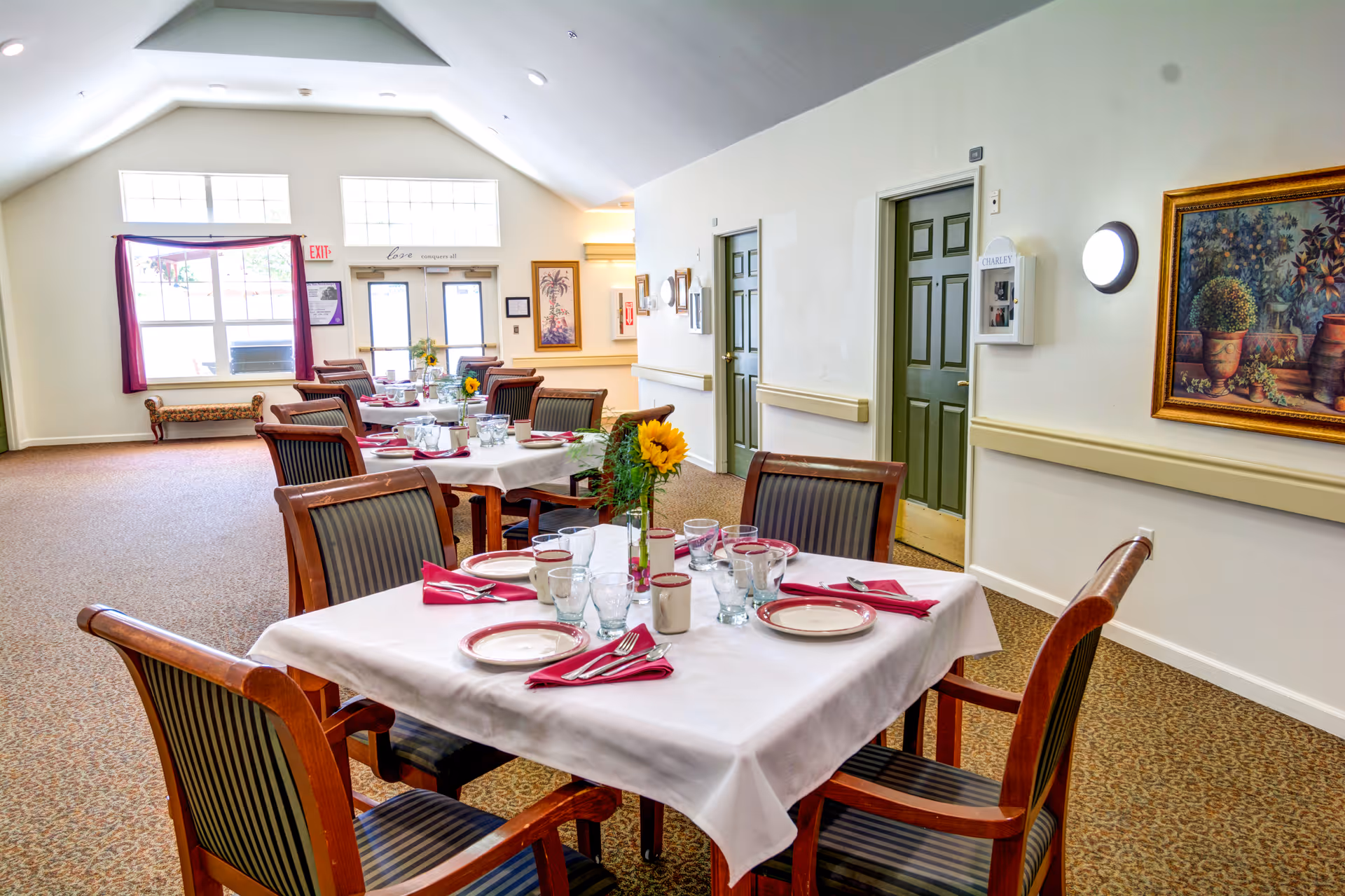 A dining room in Callahan Court Memory Care with tables set for meals. Each table is covered with a white tablecloth and set with plates, glasses, silverware, and red napkins. There are wooden chairs with striped cushions around the tables. The room has carpeted floors, light-colored walls, green doors, framed artwork, and large windows letting in natural light.