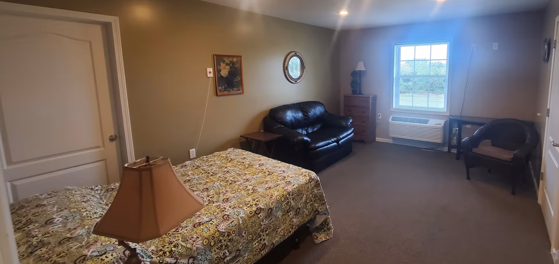 A residential care room with a bed covered in a patterned bedspread, a bedside lamp, a black leather loveseat, a wooden dresser with a lamp and decorative item, a wicker chair, a window with blinds, and neutral-colored walls with a framed picture and a round mirror.