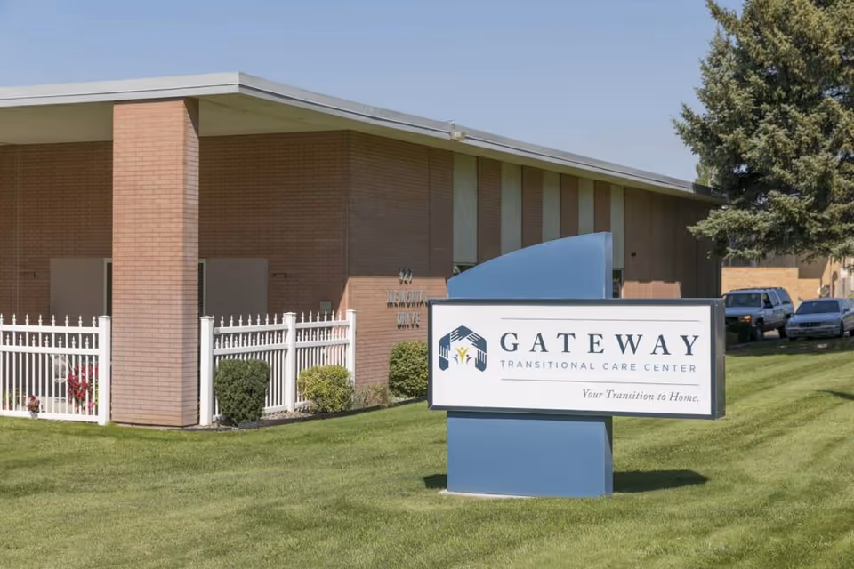 Exterior view of Gateway Transitional Care Center building with a large sign in front on a well-maintained lawn. The building is made of brick with a white fence and some bushes near the entrance. Several parked cars and a tree are visible in the background.