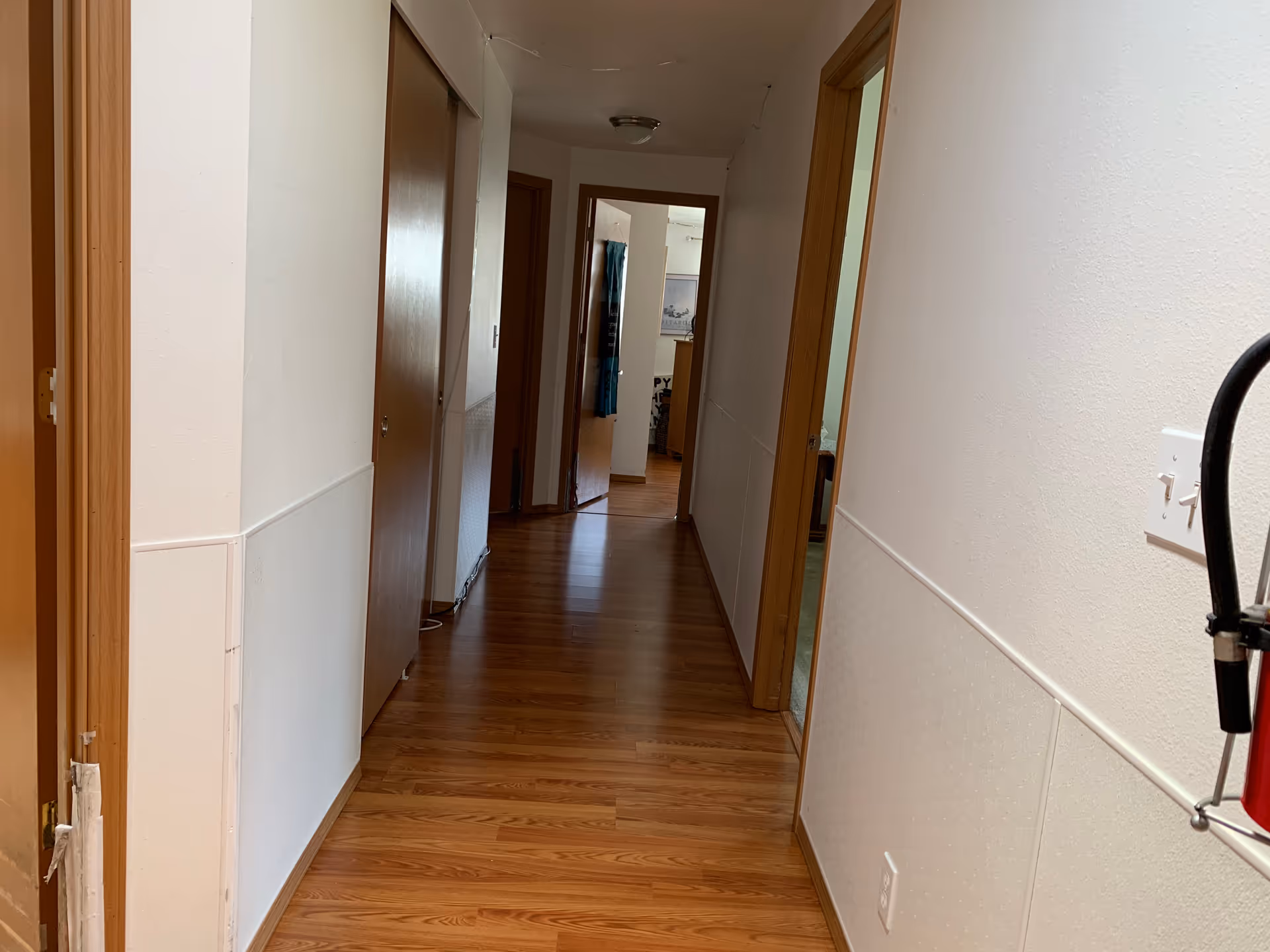 A hallway with wooden flooring and white walls, featuring several wooden doors on both sides. A fire extinguisher is partially visible on the right side near the foreground. The hallway leads to a room with a window covered by a blue curtain.