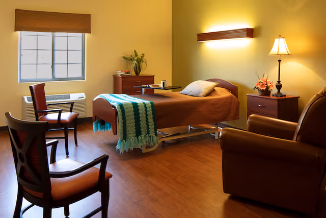 Well-lit nursing facility bedroom with a single adjustable bed, chairs, a recliner, nightstands, lamp, and a window.
