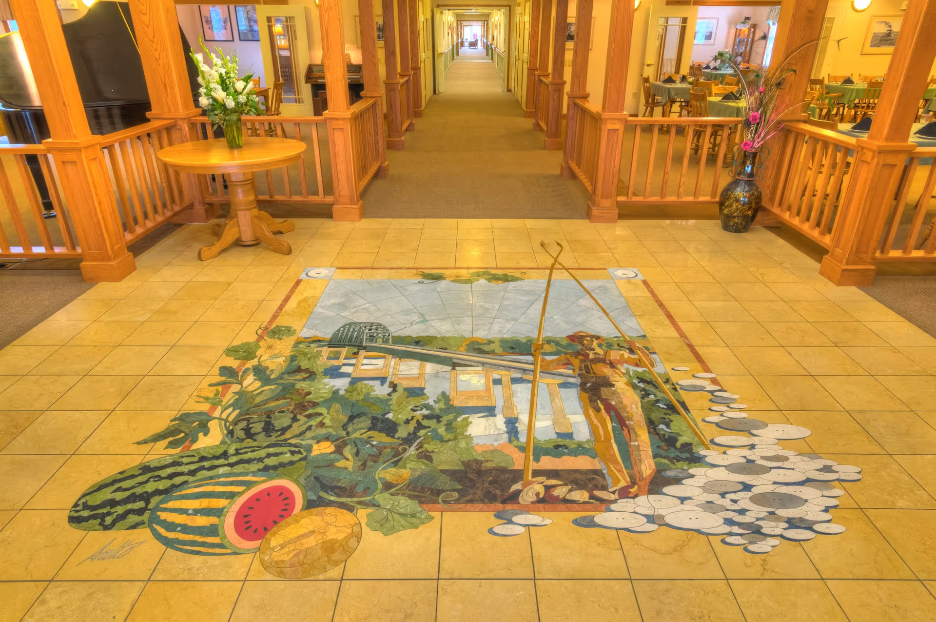 A spacious hallway in a senior living facility with wooden railings and columns. The floor features a large colorful tile mural depicting a person fishing near a bridge with watermelons and other produce in the foreground. There is a round wooden table with a vase of white flowers on the left and a large decorative vase with flowers on the right. The hallway extends into the distance with rooms and seating areas visible on both sides.