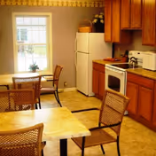 A small kitchen and dining area with wooden cabinets, a white refrigerator, a white stove, a microwave, and a kettle. There are two tables with wicker chairs around them, and a window with a view outside. The walls are painted light purple with a decorative border near the ceiling.