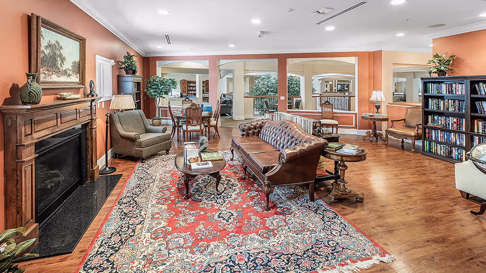 A cozy senior living common area with a brown leather sofa, an armchair, a wooden coffee table with books, a fireplace with a painting above it, a large patterned rug, wooden flooring, bookshelves filled with books, and several chairs and lamps. The walls are painted in warm tones with large windows and mirrors reflecting the space.