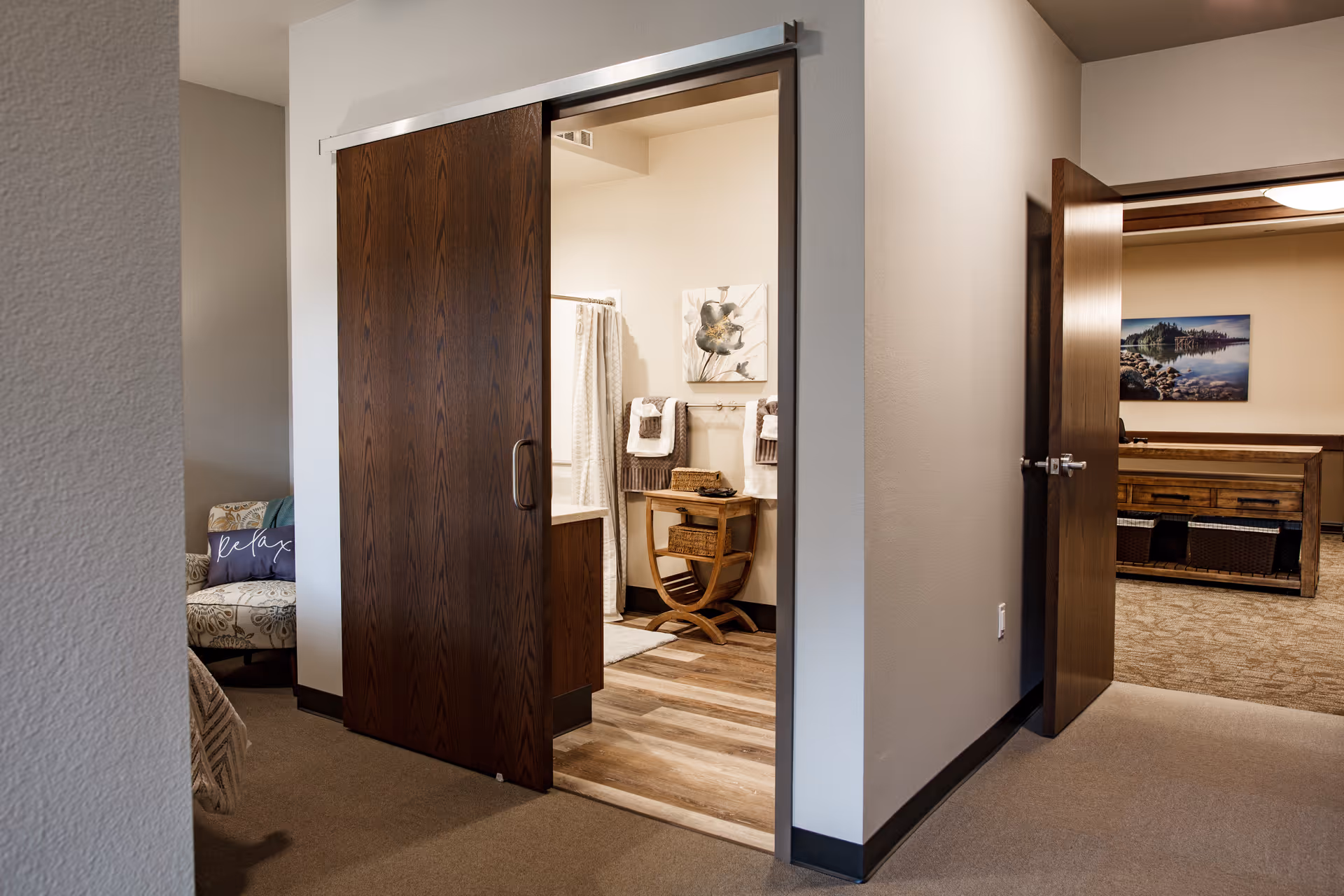 View of an interior hallway in a senior living facility showing a partially open dark wooden sliding door leading to a bathroom with towels and a small wooden table. To the right, an open door reveals a room with a wooden table and a landscape picture on the wall. A cushioned chair with a pillow that says 'Relax' is partially visible on the left.