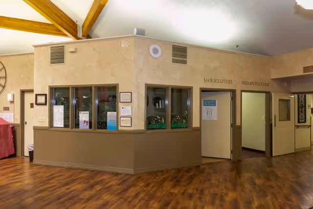 Interior view of a senior living facility hallway with wood flooring and beige walls. There is a reception or office area with windows and several certificates or notices displayed on the wall. Two doorways are labeled 'Hair Sculpture' and 'Resident's Lounge'. The ceiling has exposed wooden beams and recessed lighting.