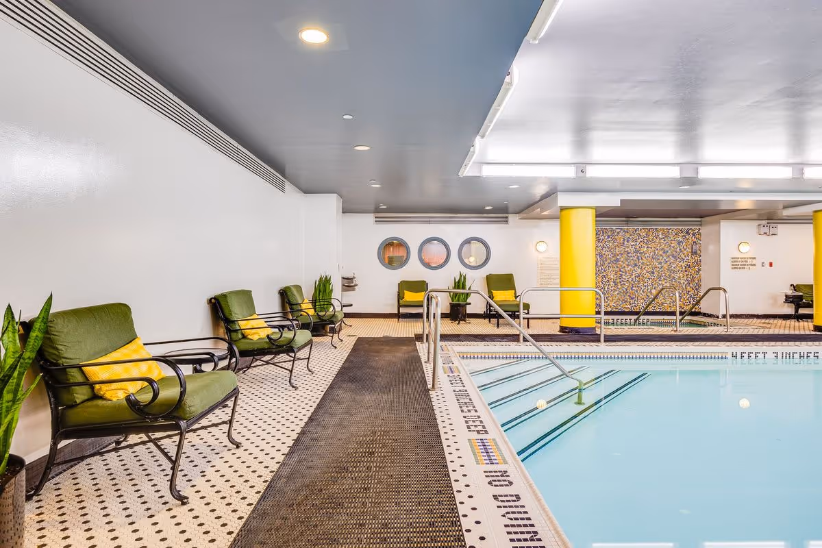 Indoor swimming pool area with a tiled floor and a row of green cushioned chairs with yellow pillows along the left side. The pool has metal handrails and depth markings on the edge. The ceiling is gray with recessed lighting, and there are three round windows on the far wall with some potted plants near the chairs.