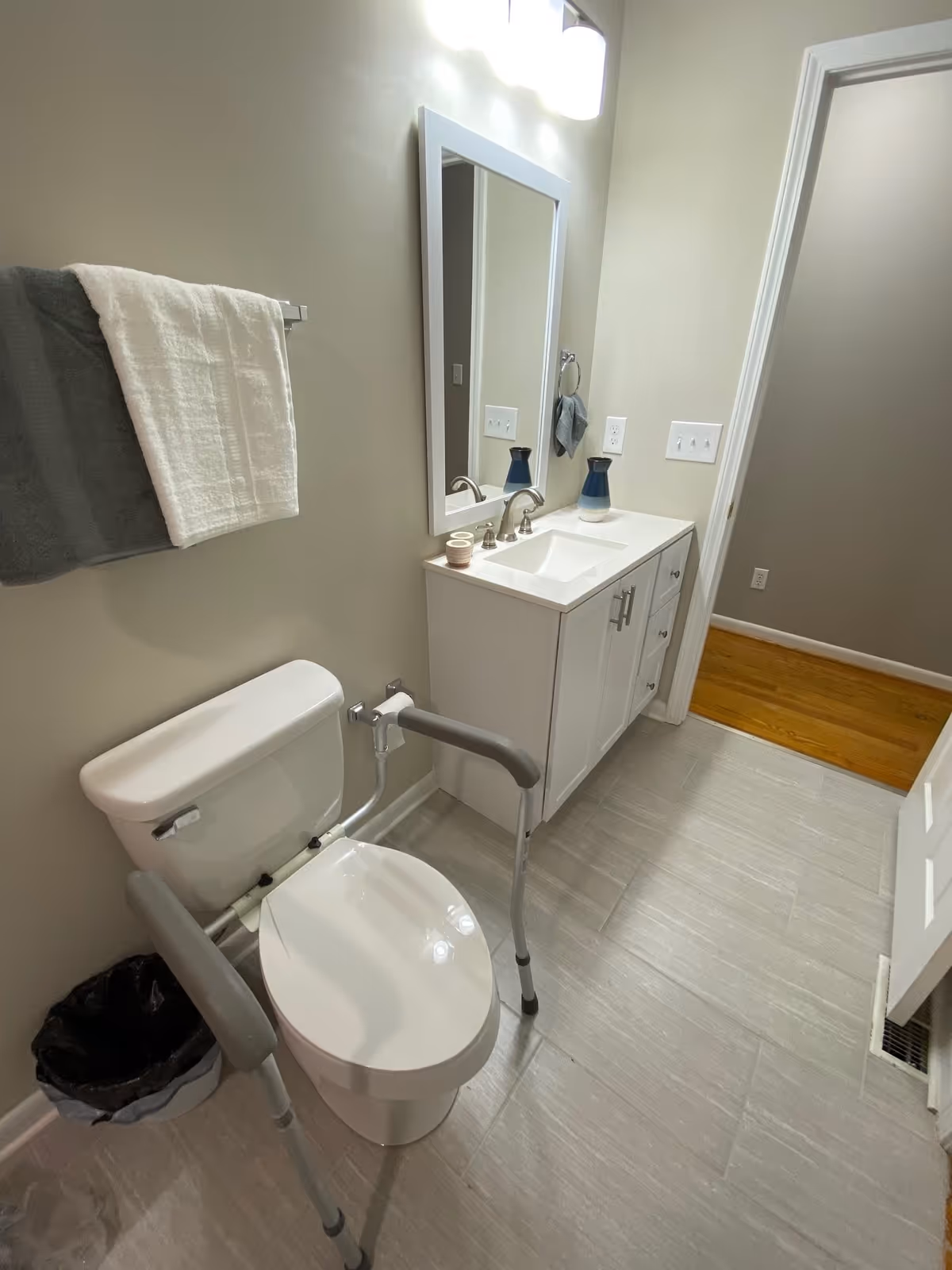 A clean and accessible bathroom with a white toilet equipped with safety rails, a white vanity with a sink, a mirror above the sink, a towel rack with a white and gray towel, and light gray tiled floor. The bathroom door is open, showing a wooden floor outside.