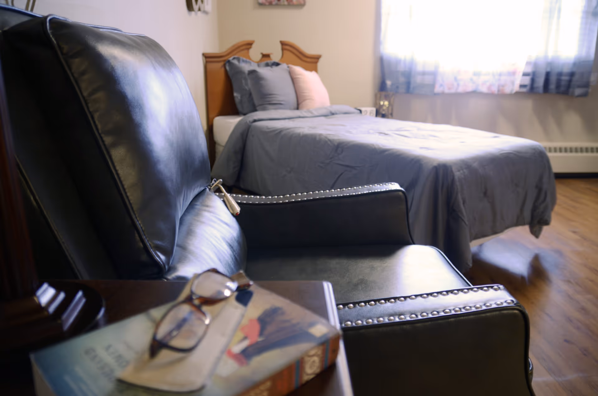 A leather armchair and side table with glasses and a book in the foreground and a made bed near a curtained window in the background.