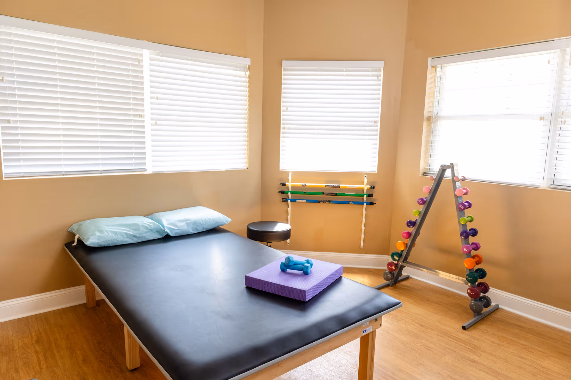 A therapy or exercise room with a black padded treatment table topped with two light blue pillows, a purple foam block, and a small blue dumbbell. The room has light brown walls, three windows with white blinds, a black round stool, a rack with colorful dumbbells, and a wall-mounted rack holding three exercise bars.